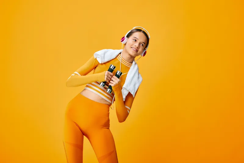 Energetic woman in bright orange workout outfit, towels over shoulder, headphones on, holding bottles, posing with motion against a vibrant orange studio backdrop for fitness and active lifestyle.