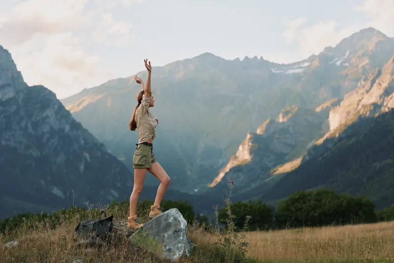 Young woman with short hair hikes in a mountain landscape, raising arms in triumph against rocky peaks and a vast blue sky