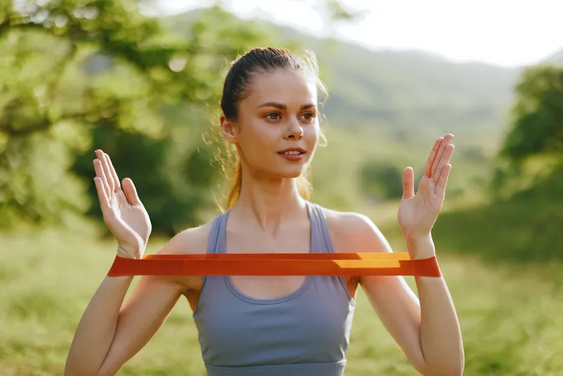 Young woman exercising with resistance band in a sunny natural landscape, promoting health and fitness in an outdoor setting