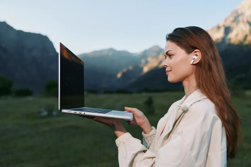 A professional woman stands outdoors with a laptop in a scenic valley. She gazes ahead with confidence, suggesting focus, productivity, and calm amid natural surroundings.