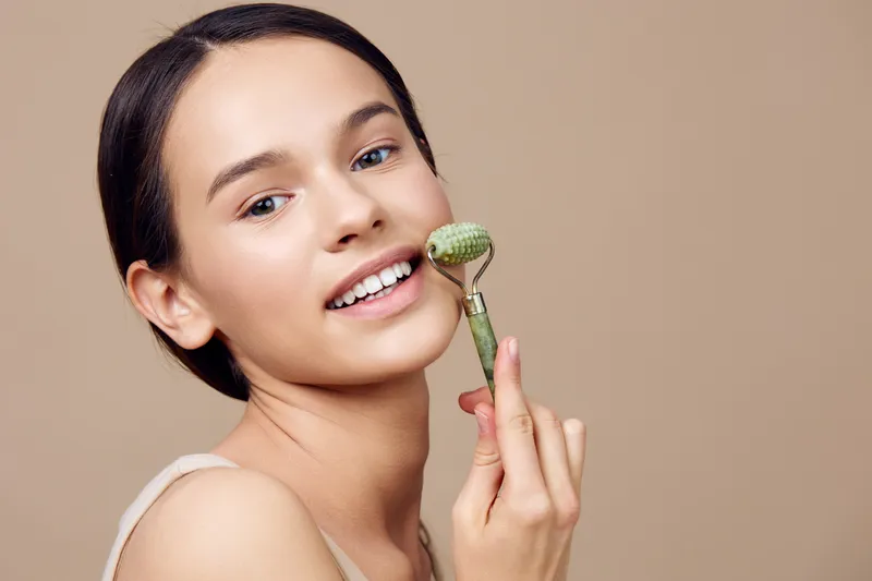 Young woman using a jade roller on her face for skincare, promoting healthy and radiant skin in a soft beige background for beauty related concepts
