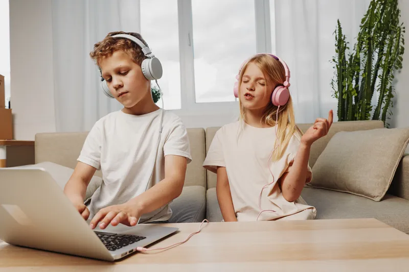 Two children wearing headphones enjoy music at home, boy using laptop and girl singing with closed eyes. Cozy living room atmosphere with natural light.