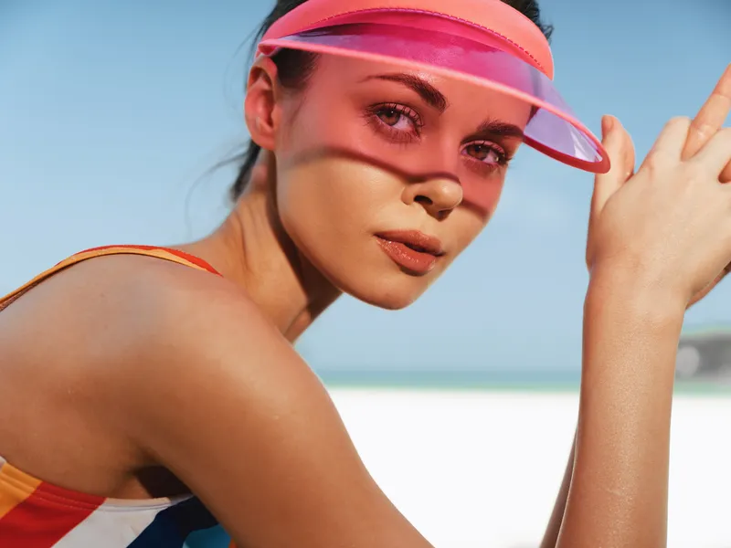 Close up of a sunlit woman at the beach wearing a colorful swimsuit and a pink visor, gazing softly toward the camera with a relaxed, confident expression.