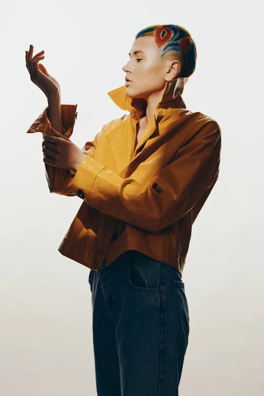 Stylish woman with colorful shaved hairstyle wearing yellow leather jacket and blue jeans poses against neutral background, showcasing modern fashion and self-expression.