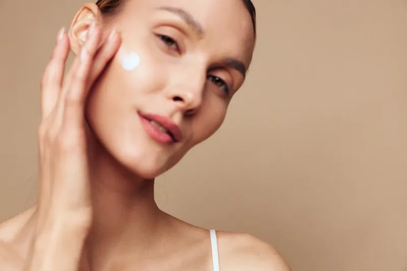 Close up portrait of a woman applying cream to her cheek, showcasing clear skin and a calm skincare routine, isolated on a neutral background, emphasizing self care and radiance