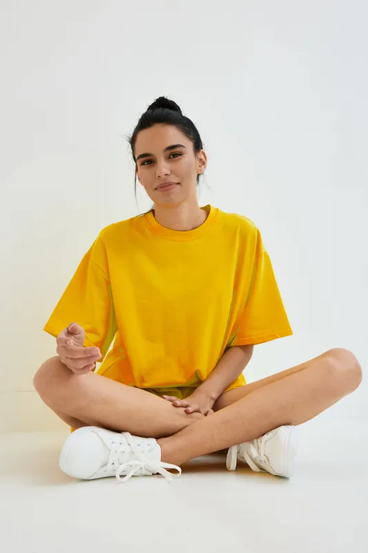 young woman sitting cross-legged indoors, wearing bright yellow t-shirt and white sneakers, relaxed expression, minimalistic white background, casual style portrait