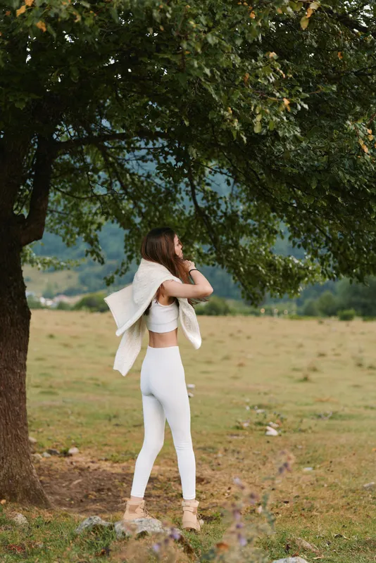 Young woman in white activewear standing under tree in nature, holding jacket on shoulders, casual lifestyle, outdoor exercise, peaceful environment