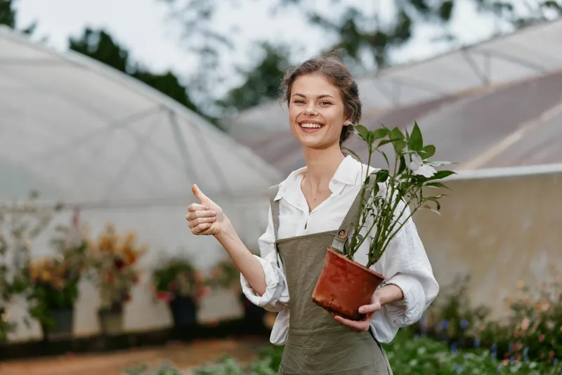 Young woman in a greenhouse giving a thumbs up, holding a potted plant, exuding joy and enthusiasm for gardening and nature