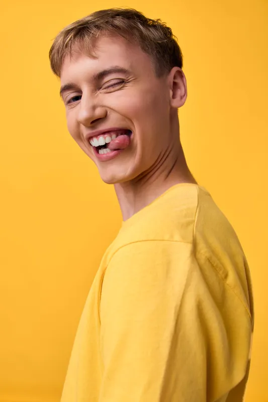 Playful expression with tongue out, yellow background, casual clothing, and a happy mood captured in a bright studio shot featuring a young man in bold color.