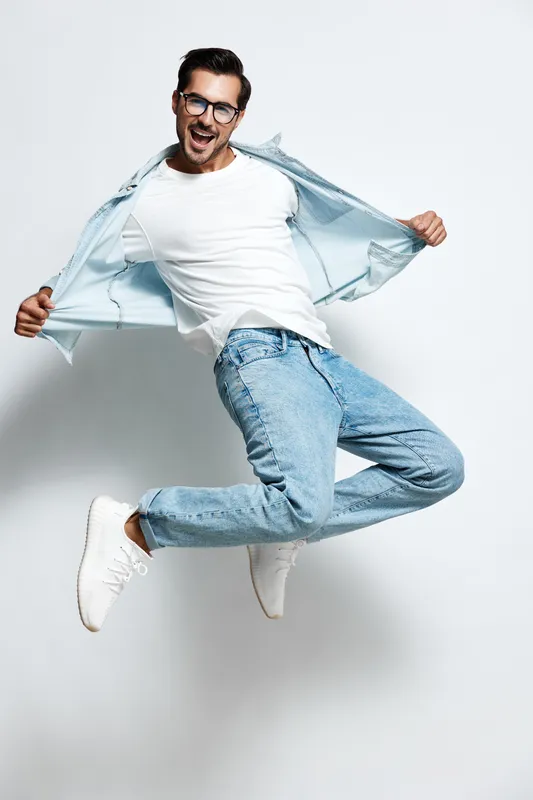 Energetic jumping happy young caucasian man wear white t-shirt and blue jeans with denim jacket isolated on plain light background studio portrait. People lifestyle concept