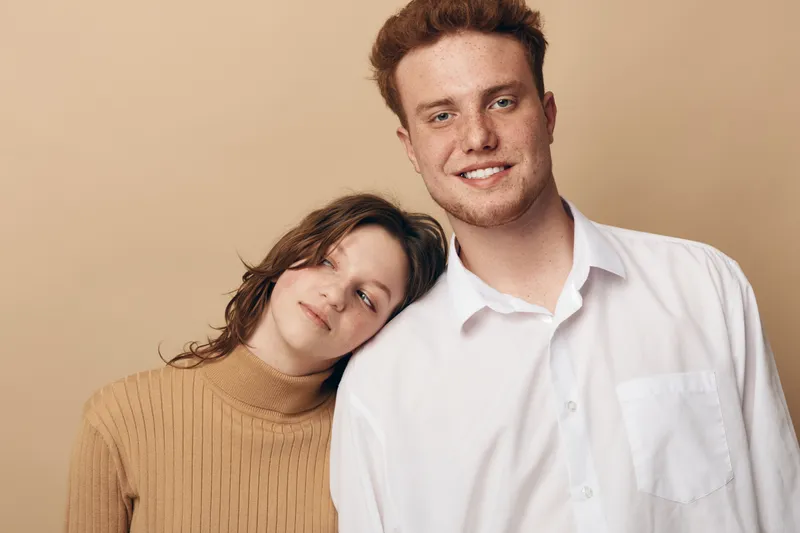 Young man and woman pose together against beige background. The woman leans her head on the man’s shoulder. Both have relaxed expressions and casual clothing. Concept of friendship and comfort.