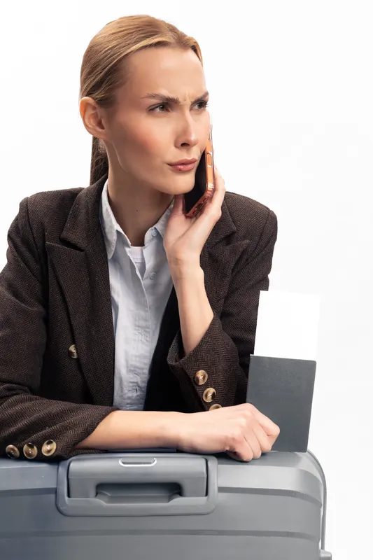 Serious businesswoman in brown blazer talking on phone while holding passport near grey suitcase. Concept for travel, business trip, communication, and planning on white background.