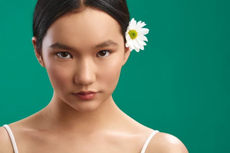 asian woman with a calm expression poses against a vivid green backdrop, showcasing natural beauty and a delicate flower behind her ear in a clean studio portrait.
