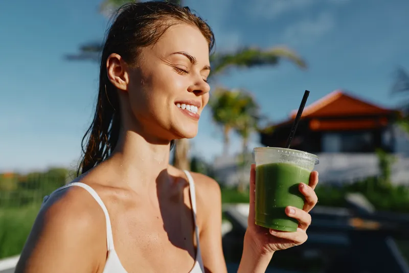 Smiling woman outdoors holding green smoothie, enjoying sunny day, relaxed posture, casual white tank top, tropical background with palm trees and blue sky