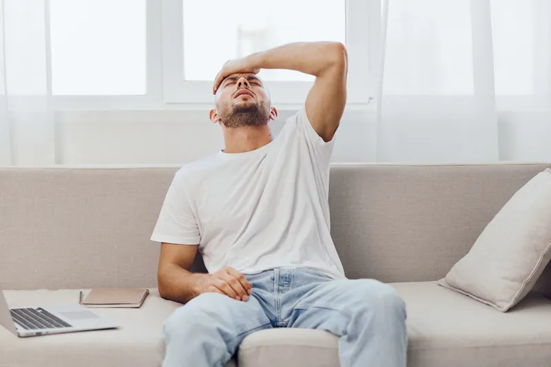 Stressed man seated on a couch, looking frustrated while holding his head. Cozy indoor setting with natural light, perfect for depicting emotional struggles.