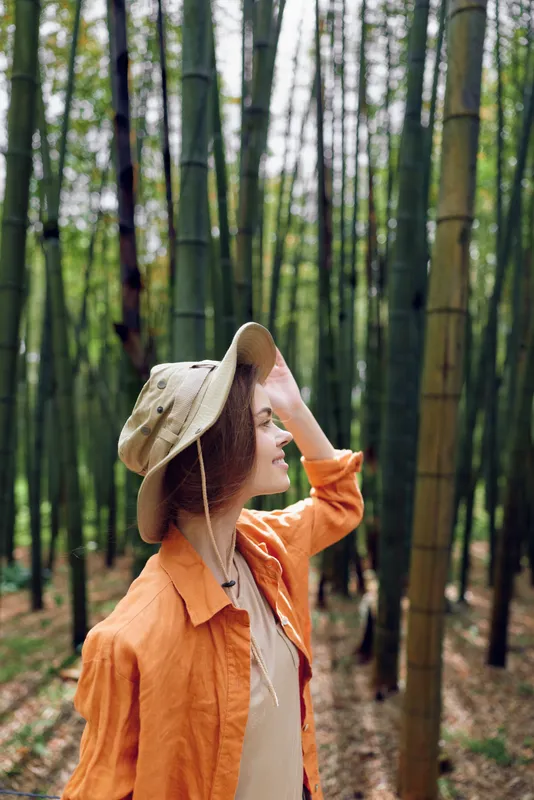 Woman in bamboo forest wearing a wide brim hat and orange shirt, smiling in profile. Portrait of nature travel and outdoor exploration with serene green background.
