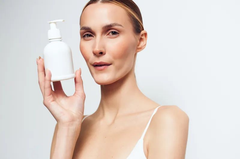 Young woman with clean skin holds white lotion bottle close to face shows skincare product for beauty routine isolated on white background with natural expression