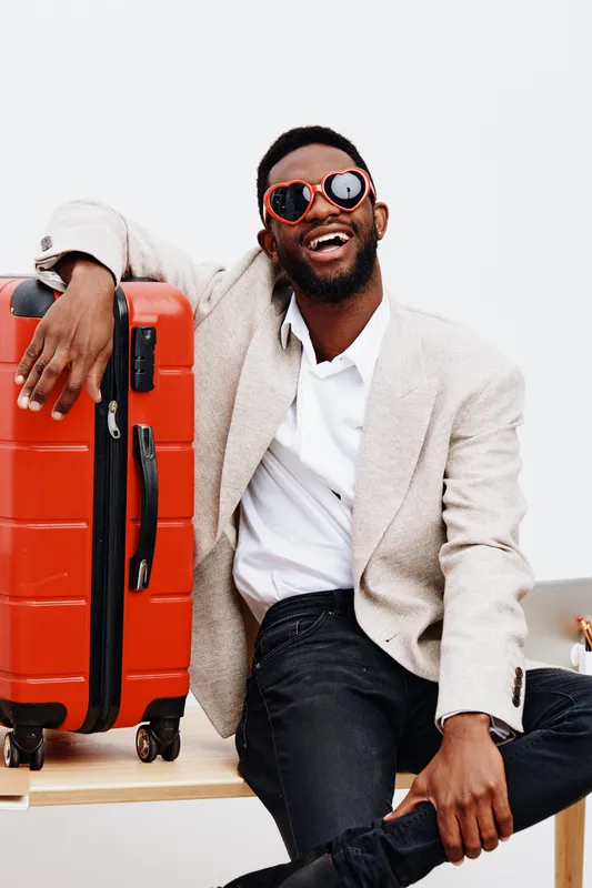 Joyful smiling man with dark skin, wearing beige blazer, white shirt, and stylish sunglasses, sitting indoors with bright background, holding orange suitcase, cheerful mood, travel concept, casual fas