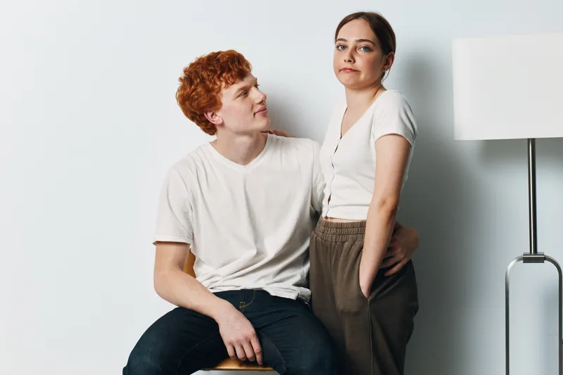 Young couple with casual clothing poses indoors against a white background, man sitting and woman standing with hands in pockets in modern studio with lamp nearby.