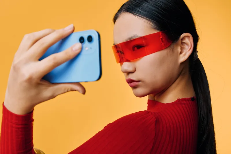 Young woman in red sweater with red futuristic glasses holding blue smartphone taking selfie on orange background. Closeup portrait of Asian female with sleek hair posing with mobile device.