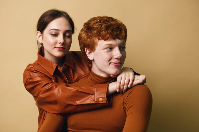 Young couple embracing with woman hugging man from behind, both dressed in brown tones with warm expressions against beige background in studio portrait.