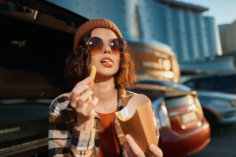 Woman eating fries by car trunk wearing sunglasses and beanie in urban parking, candid lifestyle moment with authenticity, golden hour glow and emotional storytelling, mindful living.
