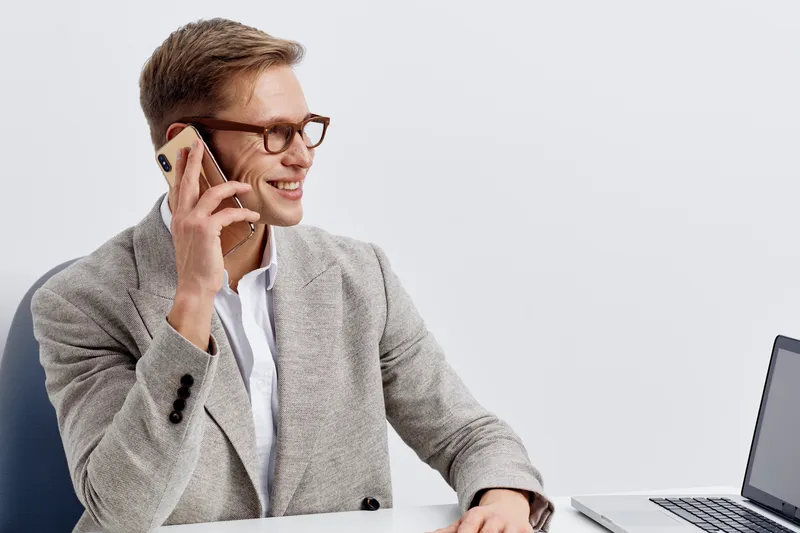 Smiling young caucasian man talking phone wearing gray blazer white shirt sitting at desk laptop digital nomad work concept communication technology