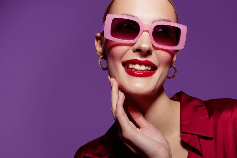Smiling woman with pink sunglasses and red lipstick poses against purple background. Fashion portrait with bright makeup and stylish accessories. Concept of beauty and confidence.