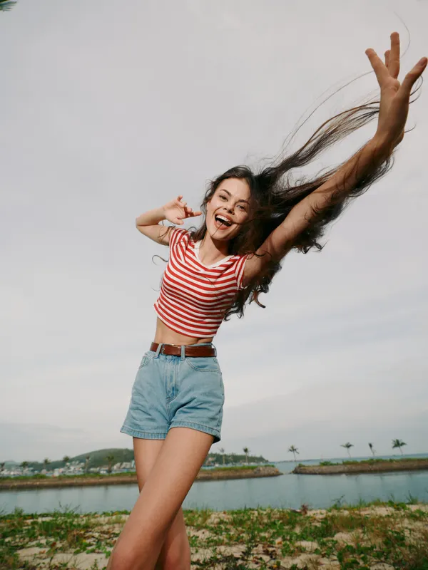 Joyful young woman jumping outdoors near a calm lake, wearing a striped crop top and denim shorts, capturing candid movement and sunny summer energy.
