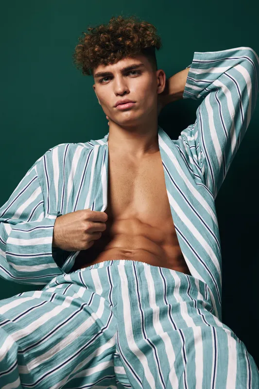 Young man with curly hair wearing striped pajamas posing against dark background, showing confident expression and relaxed posture with hand behind head.