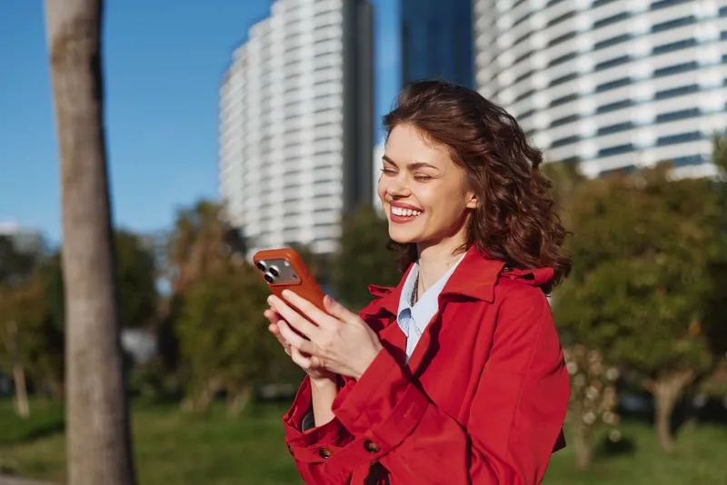 Young woman wearing a red trench coat smiles while looking at her smartphone in a sunny park, presenting a candid cheerful moment outdoors.