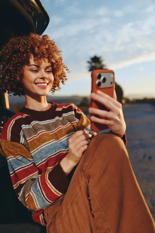 Woman lifestyle portrait outdoors at sunset, curly hair smiling, holding smartphone for selfie, wearing striped sweater and brown pants, warm evening light, relaxed casual fashion