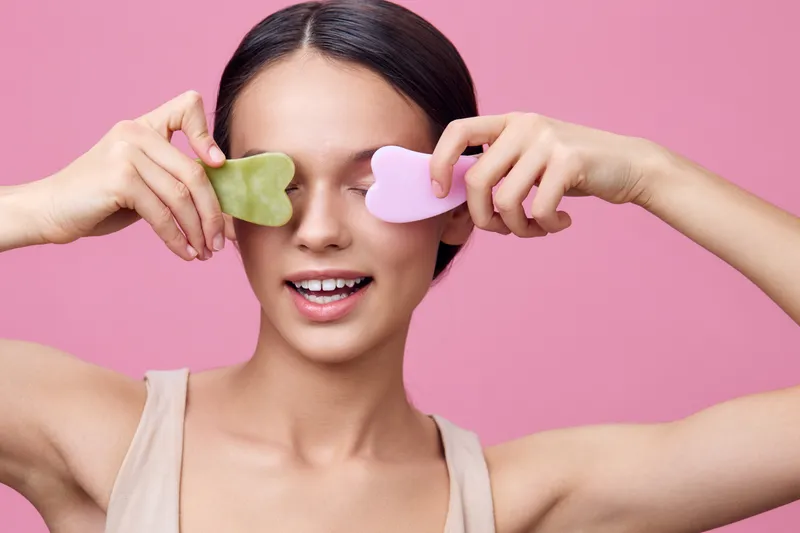 Young woman using jade rollers on her face, smiling joyfully against a pink background, promoting skincare and self care routines