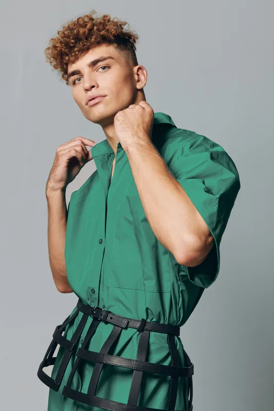 Young man with curly hair wearing transparent black belt and oversized green shirt adjusting collar on gray background. Fashion model in casual urban style with confident expression standing indoor.
