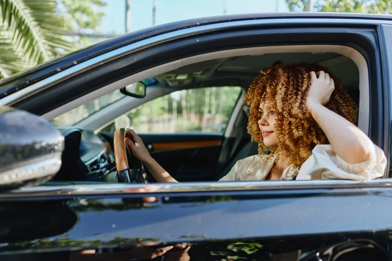 Curly woman driving a black car, smiling happily with casual outfit and natural sunlight, relaxing outdoors on a sunny day during spring season.