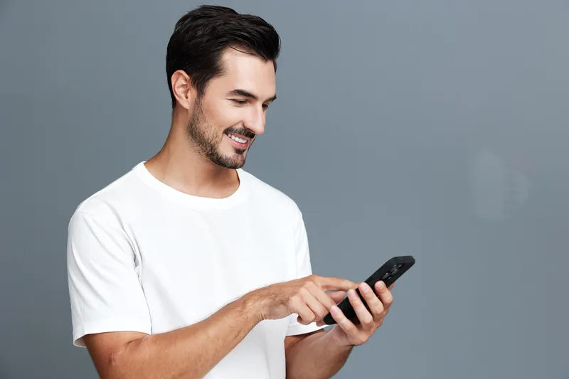 Smiling man in casual white t-shirt interacts with smartphone, expressing happiness while standing against a grey background. Modern lifestyle concept.