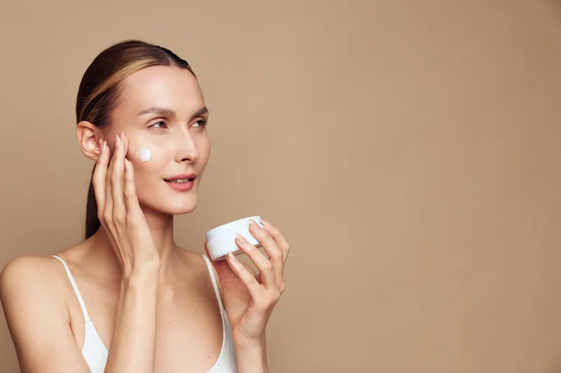 Young woman applies face cream with fingers holds jar of moisturizer has smooth skin and natural makeup isolated on beige background in skincare beauty routine concept.