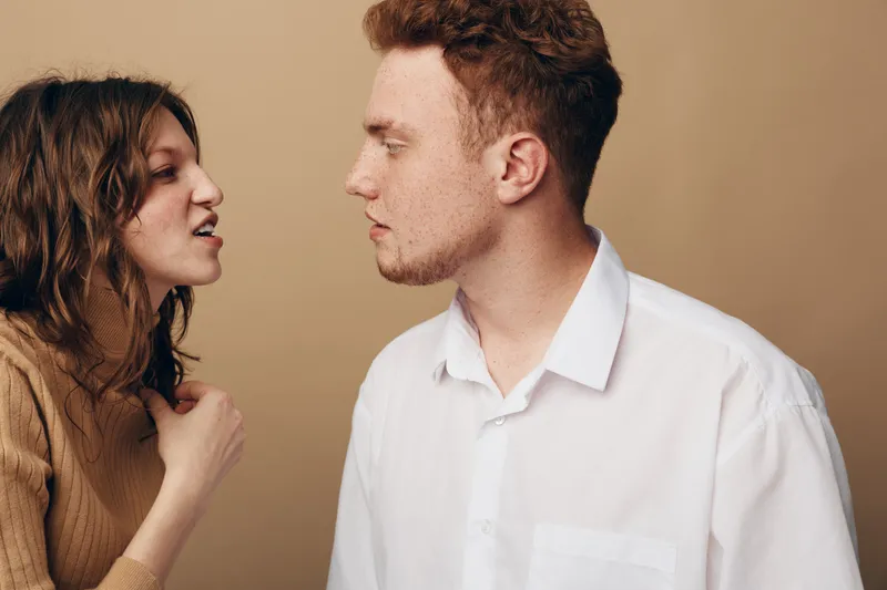 Young woman argues with young man in white shirt against beige background. Emotional conflict, disagreement, and tense communication between couple in casual clothing. Relationship tension concept.