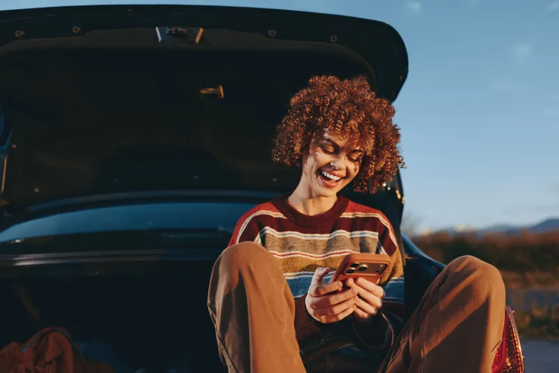 Woman lifestyle, smartphone outdoors, smiling with curly hair near car door, warm sunset light, relaxed travel moment, casual sweater, open trunk view, natural colors