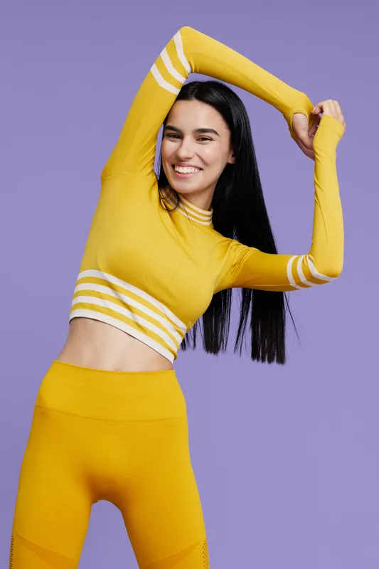 Woman sport fitness stretching in yellow activewear with a joyful smile, long dark hair, and a vibrant purple background for energetic wellness and health.