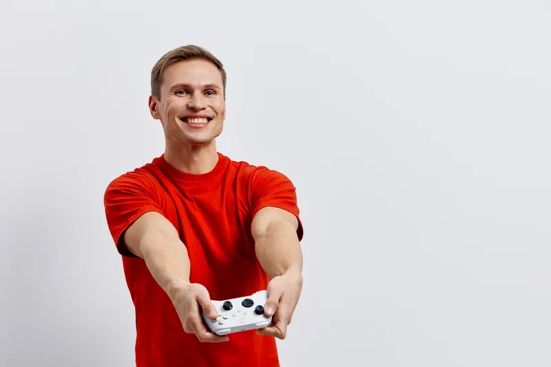 Smiling young man in red t-shirt holding gaming console and pointing forward, casual indoors, cheerful facial expression, youth lifestyle, digital entertainment, playful mood, bright lighting.