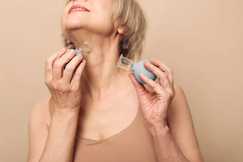 Senior woman using facial cupping therapy on neck with silicone cups in neutral studio. Concept of natural skincare, anti aging, wellness, and holistic beauty treatment.