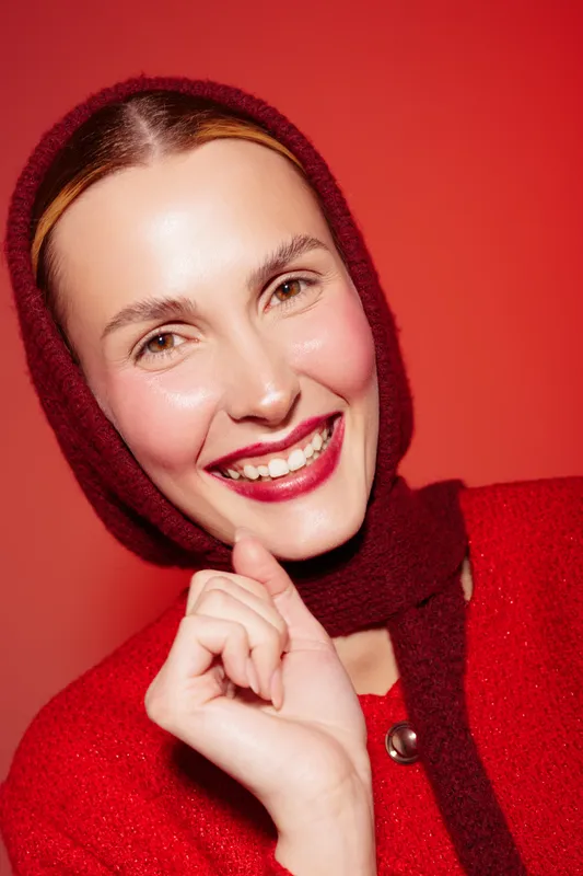 Smiling woman with red scarf and red sweater poses against red background. Close-up portrait with natural makeup and bright lipstick. Concept of fashion, beauty, and winter style.