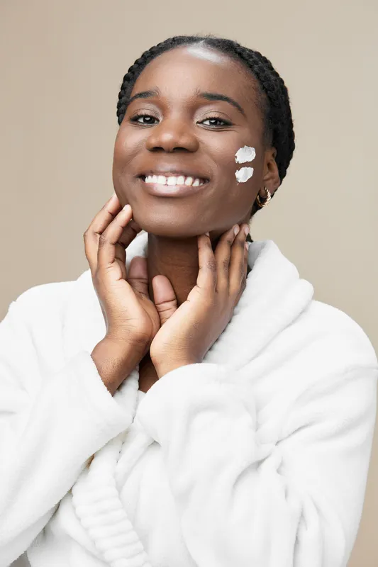 Young African woman applying skincare product with a bright smile, wearing a cozy white bathrobe against a neutral background, emphasizing self care and beauty