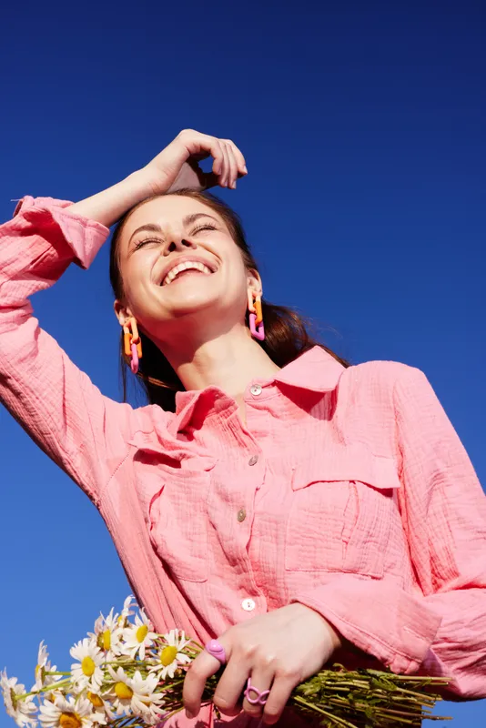 Cheerful young woman in a pink shirt holding flowers against a clear blue sky, radiating joy and vitality, perfect for spring and summer concepts