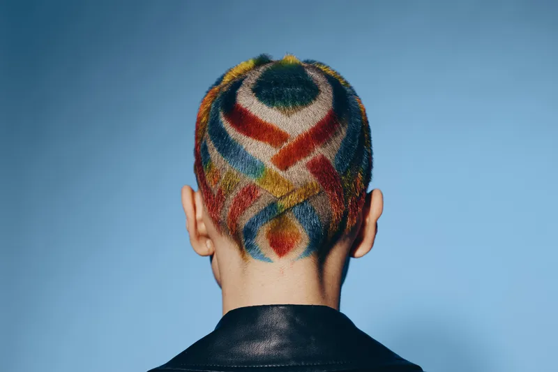 woman with colorful shaved hair art showing geometric shapes on blue background wearing black leather jacket, creative and bold hairstyle, rear view portrait