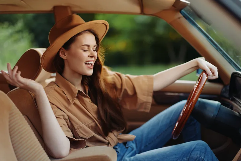 Joyful young woman in a stylish hat enjoys a carefree drive in a vintage car, showcasing a relaxed and confident vibe with a soft green background.