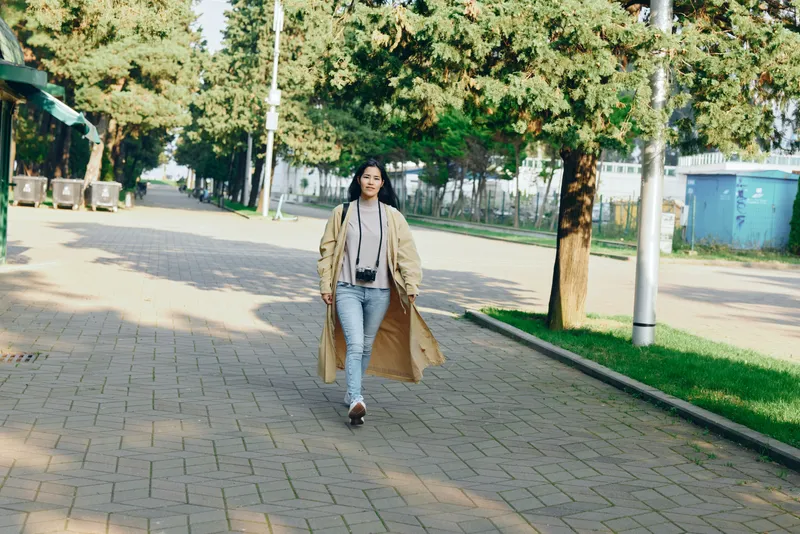 Urban street scene features a woman walking along a paved path in a long beige coat, denim jeans and boots, daylight filtering through trees, a calm stylish outdoor moment