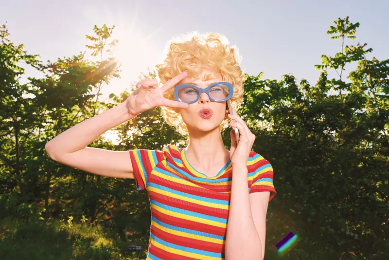 A playful woman in a striped rainbow shirt and retro glasses outdoors, sunny backdrop. A candid lifestyle portrait capturing vibrant energy and carefree summer vibe.