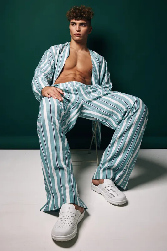 Elegant man posing sitting in relaxed striped loose clothing with open shirt and white slip on shoes against neutral studio backdrop porcelain male model fashion style.